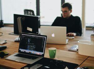 This image portrays a man working at a table. He has an open laptop and a monitor in front of him. There is another open laptop in front of him which indicates that there is another person working at the table.