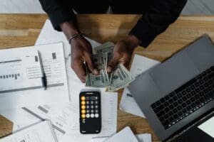 This image depicts a man putting a budget together. It features an image of a man's hands counting money at a desk that has some documents, a phone with the calculator app open and an open laptop on top of it.