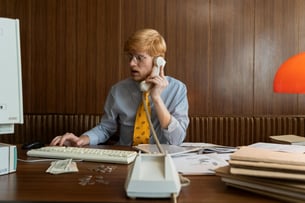 This is an image of a man working on an old computer while talking on a landline.