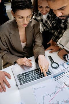 This is an image of people gathered around a desk looking at a laptop screen with documents on the desk.